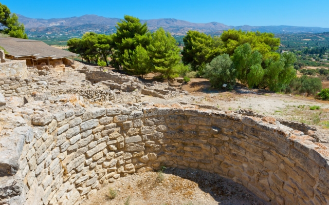 The archaeological ruins of the Palace of Phaistos on Crete, showing the well-preserved grand staircase, courtyards, and remnants of Minoan architecture, set against a backdrop of rolling hills under a clear sky.