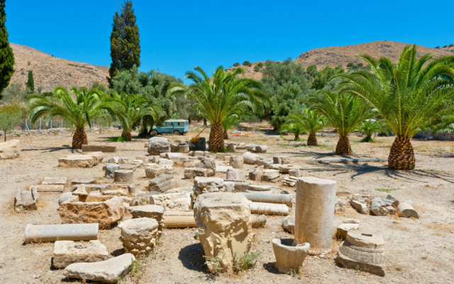 Extensive archaeological ruins of Gortys, Crete, showing remnants of Roman structures, including columns, foundations, and the Odeon, surrounded by olive groves under a clear sky.
