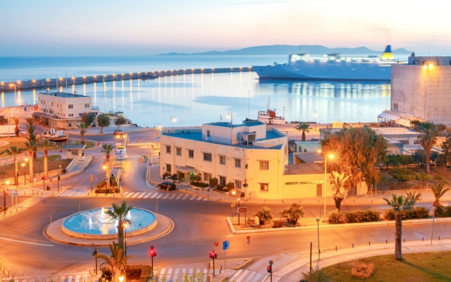 A panoramic view of Heraklion's historic Venetian harbor at sunrise, with the Koules Fortress prominently featured, fishing boats docked in the calm waters, and the sky displaying soft, warm colors.