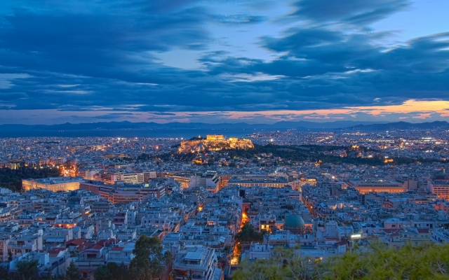 A majestic view of the Acropolis in Athens, Greece, at twilight, with the Parthenon and other ancient structures beautifully illuminated against a deep blue and orange sky, overlooking the city lights.