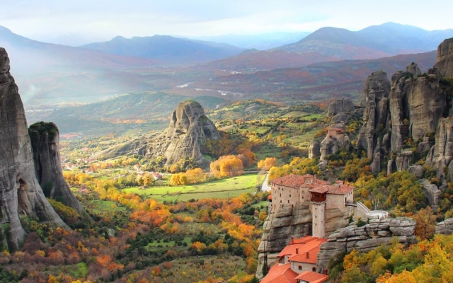 A panoramic view of the Meteora region in Greece, featuring ancient monasteries dramatically built atop towering, sheer rock pinnacles, surrounded by a lush green landscape under a vast sky.