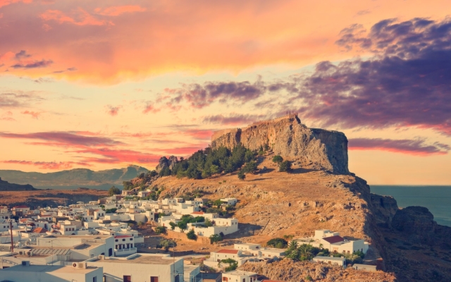 A panoramic view of the whitewashed village of Lindos, Greece, cascading down a hillside towards the turquoise Aegean Sea, crowned by the ancient Acropolis with its Doric temple and medieval castle walls.