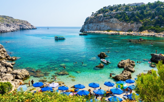 A stunning view of Anthony Quinn Bay in Rhodes, Greece, featuring emerald-green, crystal-clear waters, surrounded by rugged rocky cliffs and lush green vegetation, with small boats anchored in the bay.