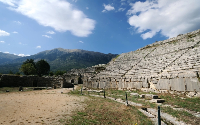 The well-preserved ruins of the ancient theater at Dodoni, Greece, with its large stone seating tiers and stage area, surrounded by lush green hills under a bright sky.
