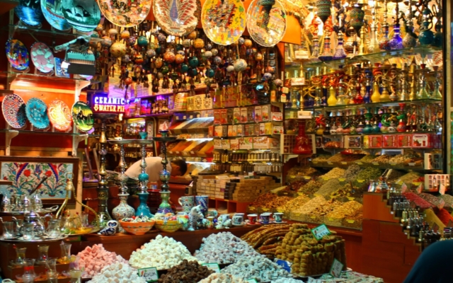 A bustling interior view of Istanbul's historic Grand Bazaar, with vaulted ceilings, colorful stalls overflowing with textiles, ceramics, lamps, and spices, and people Browse.
