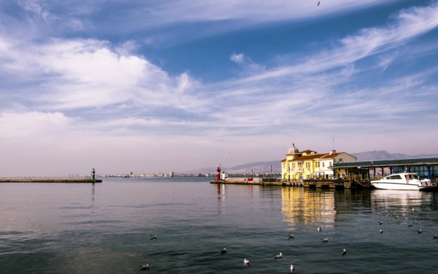 A panoramic view of Izmir's modern cityscape along the Aegean coast, with buildings extending towards the sea, a bustling promenade, and possibly palm trees and a clear sky.