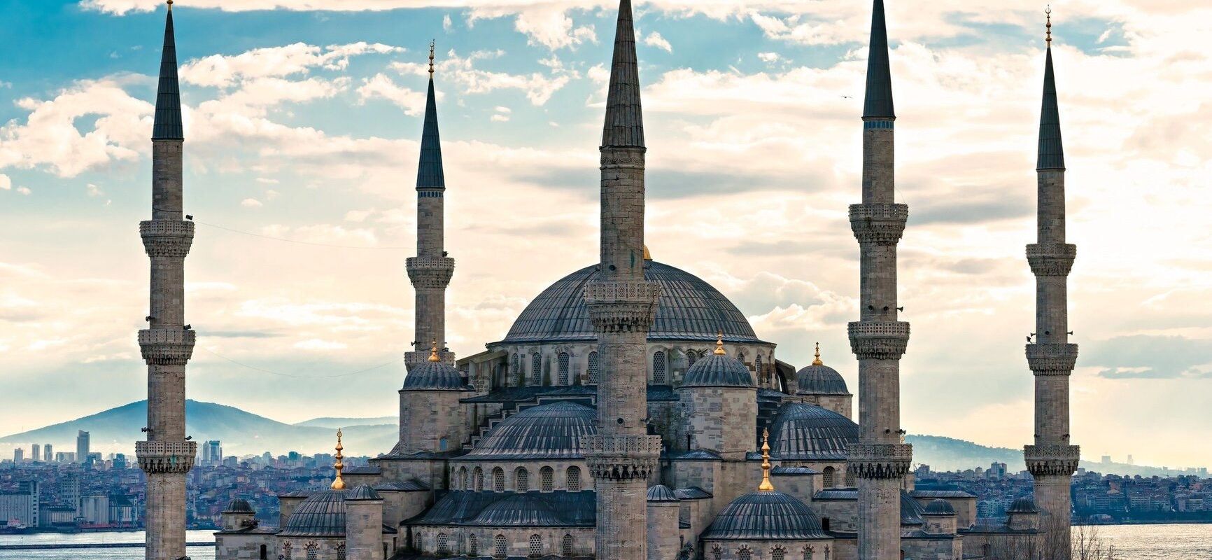 The exterior view of the historic Blue Mosque in Istanbul, with its distinctive six minarets and cascading domes, set against a clear blue sky. The intricate architecture and the surrounding courtyard are visible.