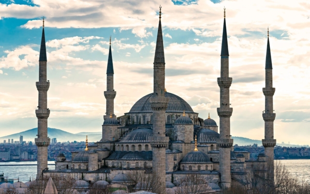 The exterior view of the historic Blue Mosque in Istanbul, with its distinctive six minarets and cascading domes, set against a clear blue sky. The intricate architecture and the surrounding courtyard are visible.