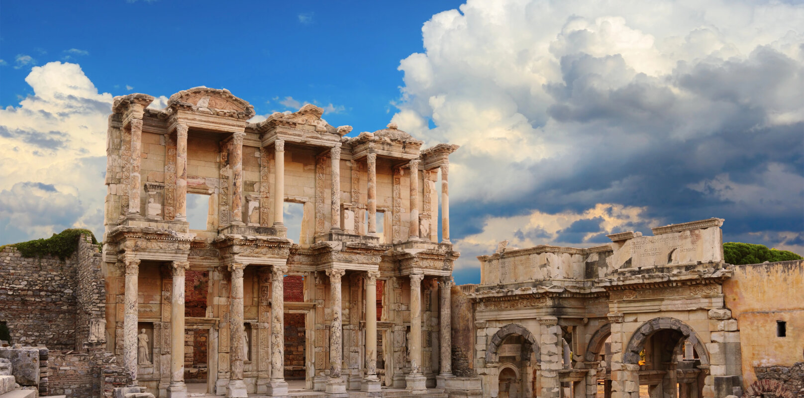The two-story facade of the ancient Celsus Library in Ephesus, Turkey, with intricate carvings, statues in niches, and a grand staircase, bathed in sunlight.