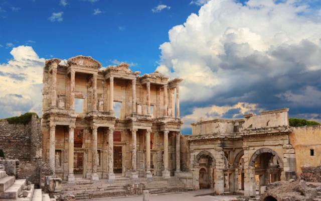 The two-story facade of the ancient Celsus Library in Ephesus, Turkey, with intricate carvings, statues in niches, and a grand staircase, bathed in sunlight.