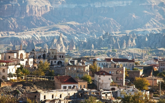 Panoramic view of Cappadocia's unique landscape, featuring towering, cone-shaped "fairy chimney" rock formations, some with cave dwellings carved into them, under a clear blue sky.