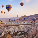 Dozens of colorful hot air balloons floating gracefully above the unique "fairy chimney" rock formations and valleys of Cappadocia, Turkey, at sunrise, with warm golden light illuminating the landscape.