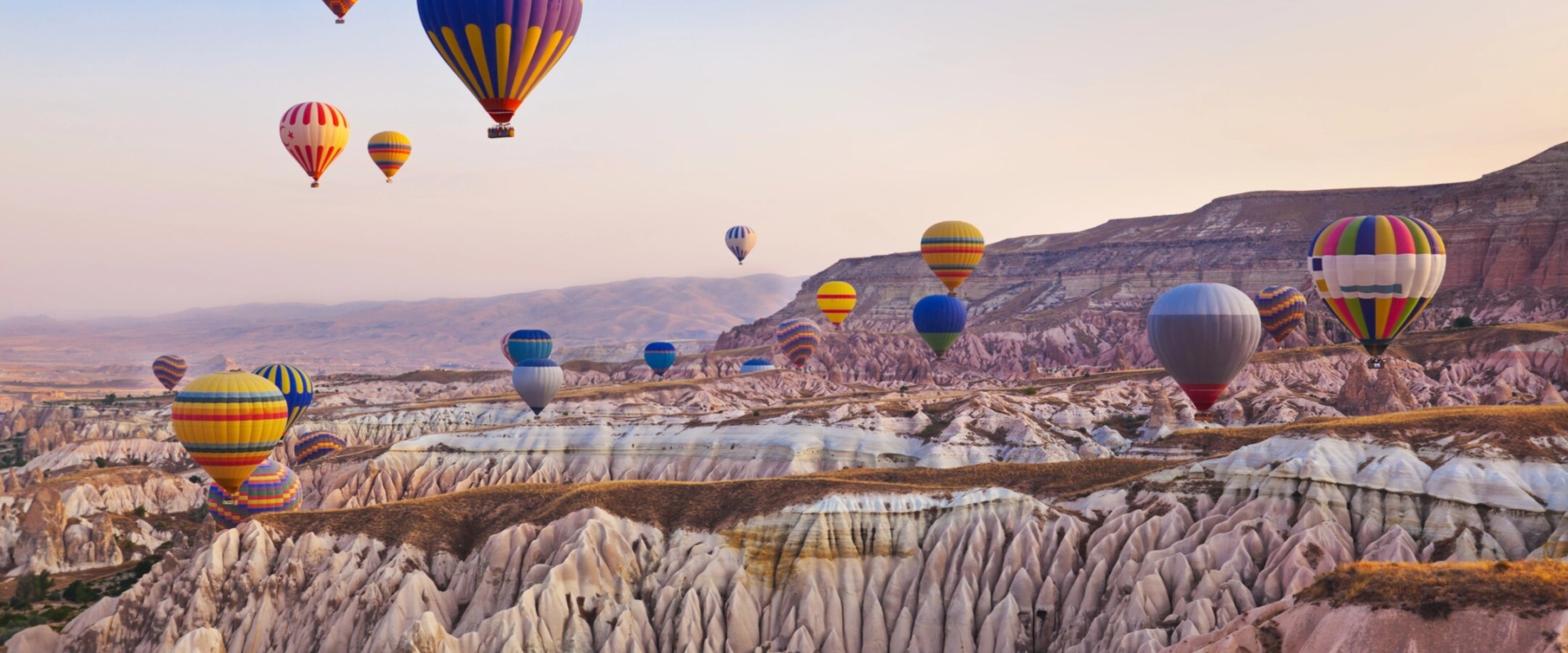 Dozens of colorful hot air balloons floating gracefully above the unique "fairy chimney" rock formations and valleys of Cappadocia, Turkey, at sunrise, with warm golden light illuminating the landscape.