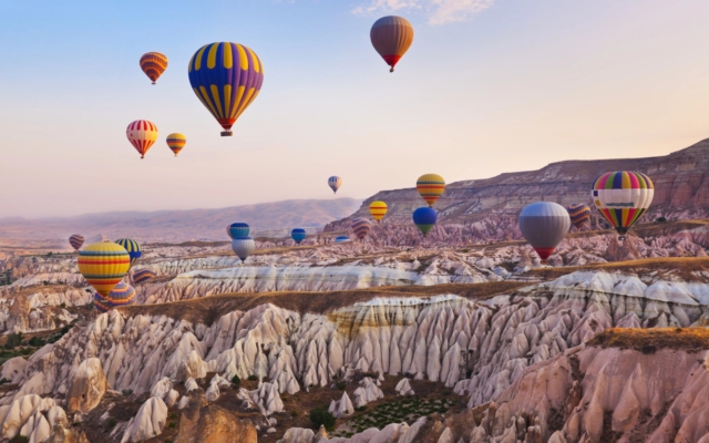 Dozens of colorful hot air balloons floating gracefully above the unique "fairy chimney" rock formations and valleys of Cappadocia, Turkey, at sunrise, with warm golden light illuminating the landscape.