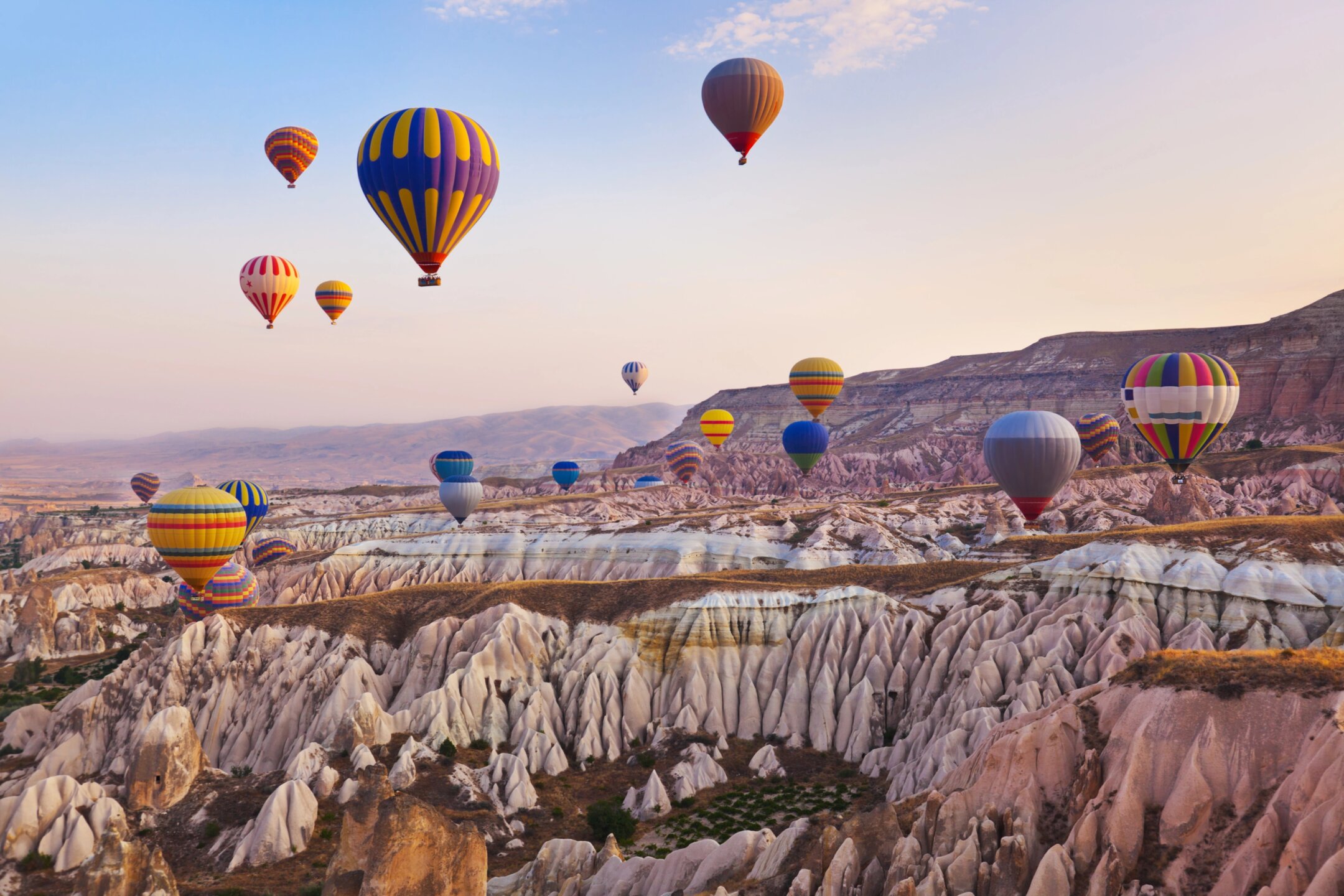Dozens of colorful hot air balloons floating gracefully above the unique "fairy chimney" rock formations and valleys of Cappadocia, Turkey, at sunrise, with warm golden light illuminating the landscape.