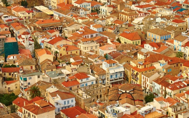 An aerial or high-angle view of Nafplio's historic town center, showing its distinctive red-tiled roofs, neoclassical buildings, narrow streets, the harbor with boats, and surrounding hills, under a bright sky.