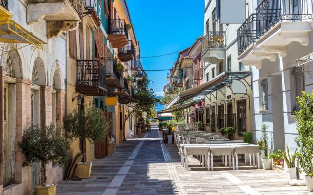 A vibrant scene of Nafplio, Greece, on a sunny summer day, showcasing its charming old town with colorful neoclassical buildings, a lively promenade along the sparkling blue sea, and boats in the harbor.